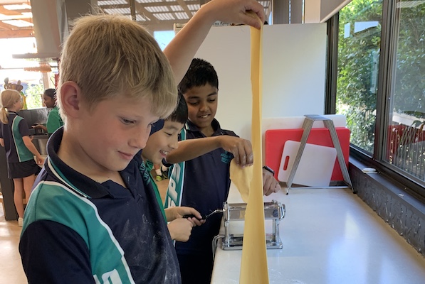 Chil pulling long strand of pasta upwards, others behind working pasta machine Chil pulling long strand of pasta upwards, others behind working pasta machine
