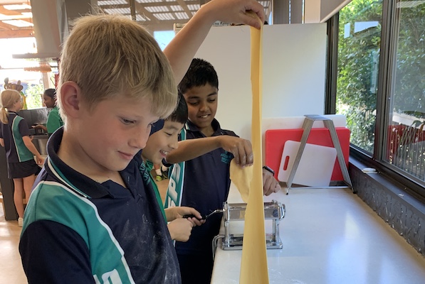 Chil pulling long strand of pasta upwards, others behind working pasta machine