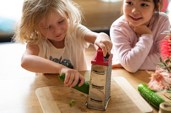 Two children, one grating cucumber while other watches on.