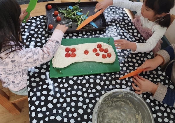 Three children placing vegetables on pizza dough.