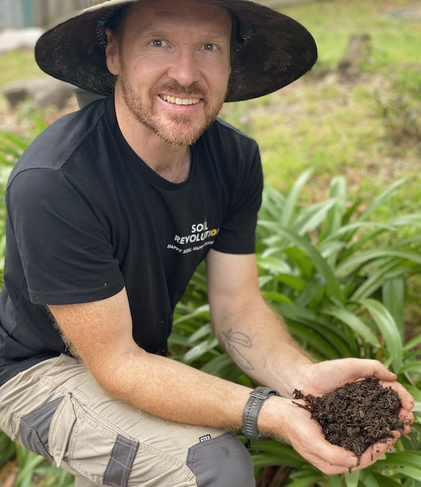 Man in wide-brimmed hat, smiling at camera, holding handful of soil and worms