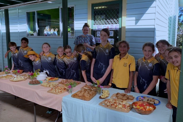 Row of children and one adult behind a table laden with dishes